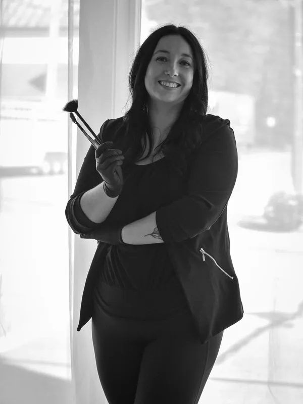 Autumn Johnson smiling and holding makeup brushes in a bright window, black and white