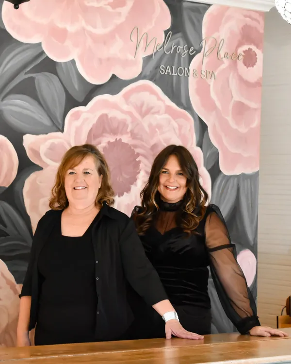 Two women smiling behind a wooden front desk, in front of the painted floral mural and gold salon sign