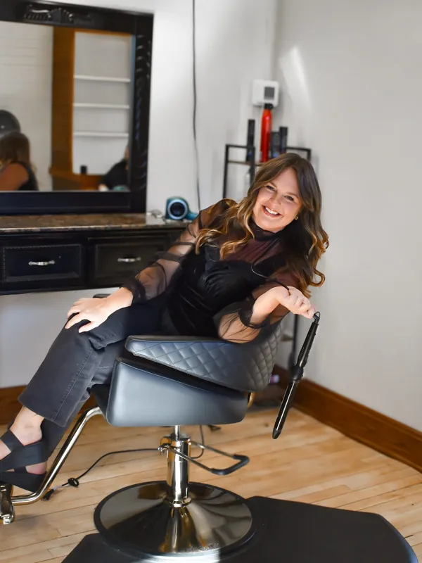Jenna Kirchner smiling in a styling chair at her station, holding a curling iron