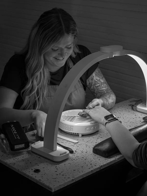 Lexi Vaningan smiling as she gives a manicure at her station under a curved lamp, black and white